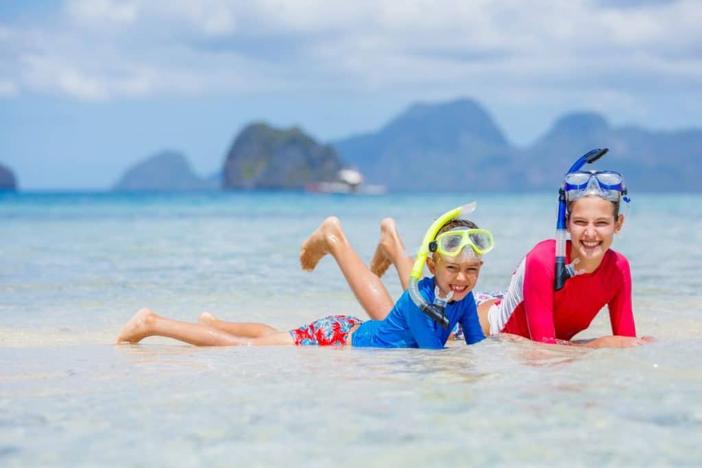 Kaksi lasta vedessä rannalla. Heillä on snorkkelit, uimapaidat ja uimahousut.